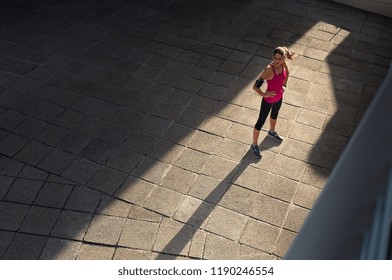 Mature Woman Taking A Break After Sport Training In City Street. Fitness Runner Relaxing After City Jogging And Working Out Outdoors. High Angle View Of Runner Standing On Sidewalk With Copy Space.