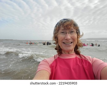 Mature woman taking a selfie before swimming in the sea. Cold or open water swimming is beneficial for both physical and mental health. She is smiling and wearing pink rash vest. Copy space on left.  - Powered by Shutterstock