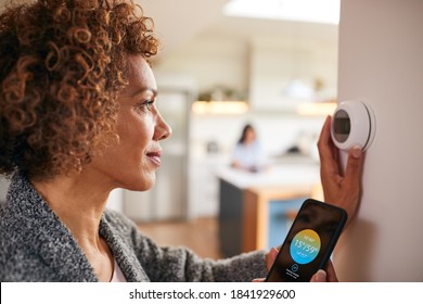 Mature Woman Using App On Phone To Control Digital Central Heating Thermostat At Home