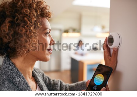 Mature Woman Using App On Phone To Control Digital Central Heating Thermostat At Home Foto stock © 