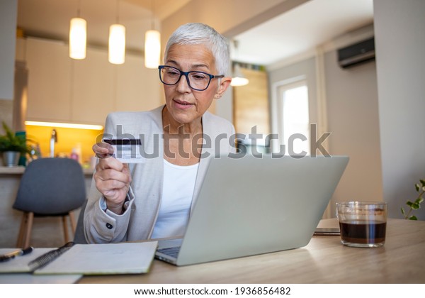 Mature woman using credit card making online payment at\
home. Successful old woman doing online shopping using laptop.\
Closeup of retired fashionable lady holding debit card for internet\
banking 