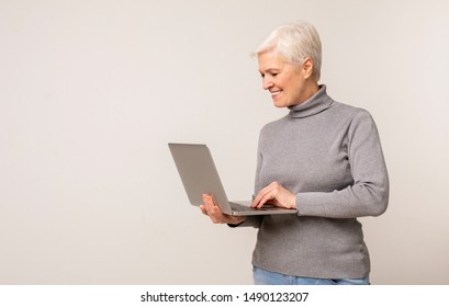 Mature Woman Using Laptop, Browsing Internet While Standing Against Light Studio Background. Copy Space