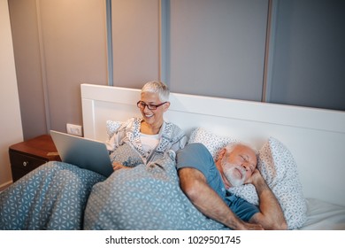 Mature woman using a laptop computer. Her husband sleeping next to her in the bedroom. - Powered by Shutterstock