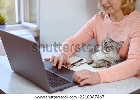 Mature woman using a laptop at home. The cat sits on her lap. There is an open notepad and a pen on the table. Online shopping, work from home, online learning and freelance concept. Focus on the cat.