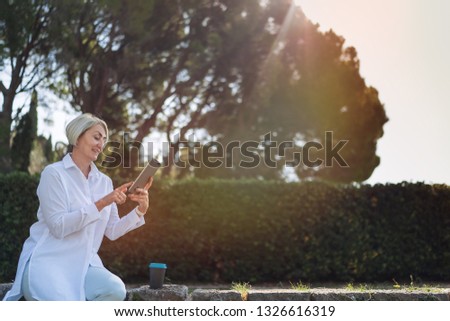 Mature woman using on tablet while resting at the park and drinking coffee