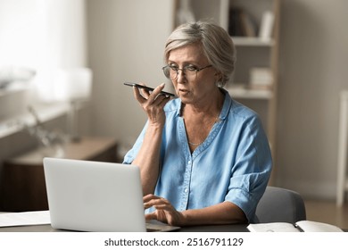 Mature woman using smartphone sit at desk with laptop, engaged in voice search or dictation on phone, input information, search data, managing work online, send audio message. Multitask, modern tech - Powered by Shutterstock