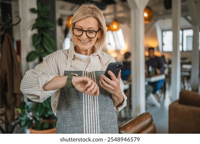 mature woman waitress stand at restaurant and use cellphone while check time on wrist watch