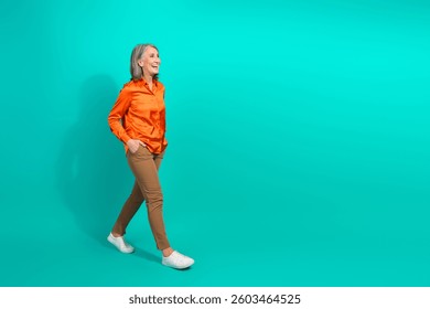 Mujer Adultas camina con confianza con una vibrante camisa naranja y pantalones marrones sobre un fondo verde azulado para fines de promoción de estilo de vida