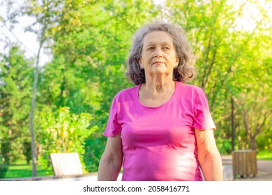 A Mature Woman Walks In The Park Against The Background Of Green Tree Crowns On A Sunny Day. An Elderly Woman With A Serious Face. Depressive Mood. Psychology. Proud Look. The Older Generation.