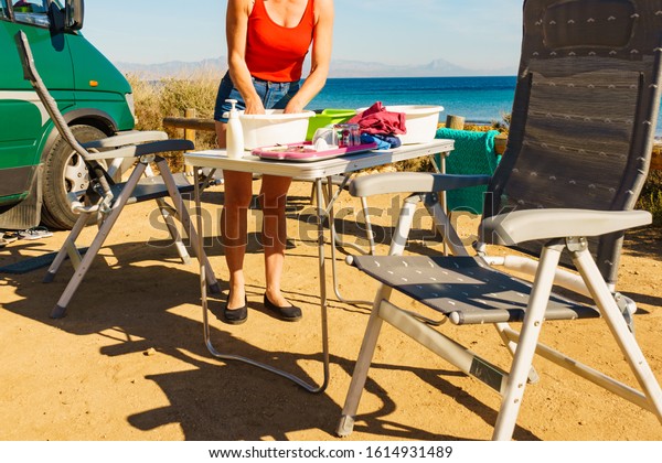 Mature\
woman washing up dishes in bowl on fresh air at camper car.\
Dishwashing outdoor on camping site, sea\
shore