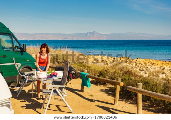 Mature\
woman washing up dishes in bowl on fresh air at camper car.\
Dishwashing outdoor on camping site, sea\
shore