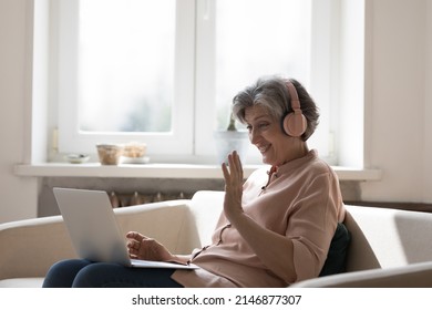 Mature Woman Wear Headphones Sit On Couch With Laptop Wave Hand Start Talk Through Videoconference App, Share News, Enjoy Remote Communication To Family Living Abroad. Virtual Meeting Event Concept