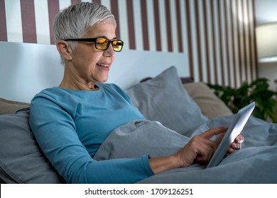 Mature Woman Wearing Blue Light Blocking Glasses With Amber Lenses, Lying In Bed Before Sleep, Looking At Tablet Screen 