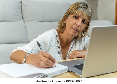 Mature Woman At Work Writing In Her Diary While Looking At The Camera And With Her Laptop Next To Her. Work From Home Concept.