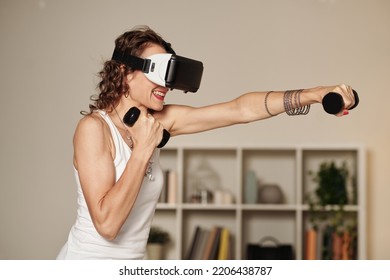 Mature Woman Working Out In Virtual Reality Headset At Home And Boxing With Dumbbells In Hands