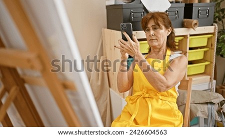 Mature woman in yellow apron photographing artwork with phone in art studio.