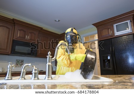 Mature woman in yellow Haz Mat suit and gas mask washing roasting pan in kitchen sink.