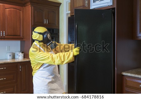 Mature woman in yellow Haz Mat sui and gas mask opening refrigerator door in home kitchen.