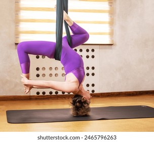 A mature yoga woman in a purple workout suit, does fly yoga and stretching, aerostretching workouts, in a hammock, the concept of Pilates therapy. Copy the space. - Powered by Shutterstock