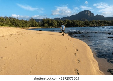 Matured caucasian man, hiker, far, walking along a sandy beach near volcanic lava rocks in the ocean and a mountain scene in the background, Mahaulepu Beach, Kauai, Hawaii - Powered by Shutterstock