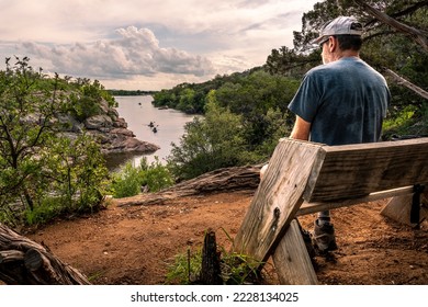 A matured Caucasion male, person, hiker, visitor, sitting on a simple wooden bench watching boats in the lake below, Inks Lake, Texas - Powered by Shutterstock