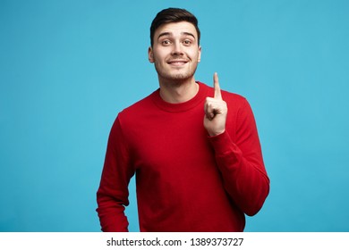 May I Have Your Attention, Please? Picture Of Emotional Unshaven Young European Guy In Red Sweater Raising Index Finger To Draw Your Attention, Going To Say Something Or Make Important Announcement