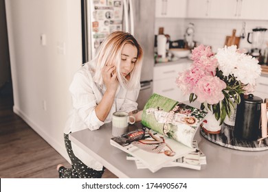 May,28,2020. Denver, United States.Young Lady Millennial.Flowers. Pajama Outfit. Bright White Kitchen. Home Lifestyle. Blonde Woman.Reading Magazines.Morning Light.Relax And Treat Yourself.Modern Life