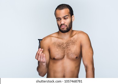 Maybe Next Time? Portrait Of Handsome Shirtless Young Black Man Looking At Razor While Standing Against White Background

