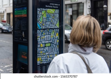 Mayfair, London/UK - November 3 2019: Young woman looks at map of the area. Selective focus on the map, which is on a black pillar. Woman has blonde hair and wearing a white jacket. Cars in background
