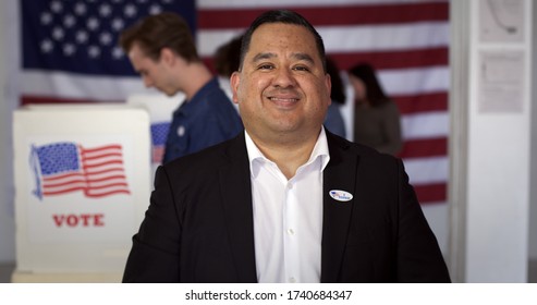 MCU Hispanic man in white shirt and black jacket with "I Voted" sticker, smiles while standing in front of other voters at polling booths with US flag in rear