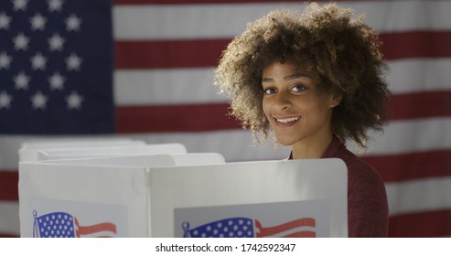 MCU Profile Young, Mixed Race Woman, Alone Casting Vote In Booth At Polling Station. She Looks Up Towards Viewer, Smiling. US Flag In Background.