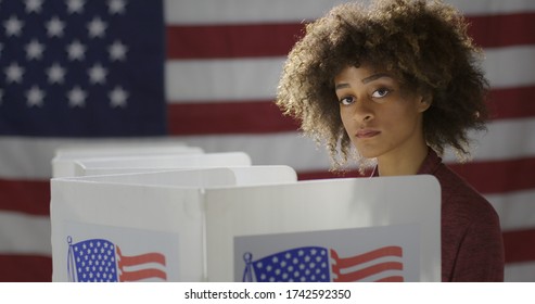 MCU Profile Young, Mixed Race Woman, Alone Casting Vote In Booth At Polling Station. She Looks Up Towards Viewer, Looking Serious. US Flag In Background.