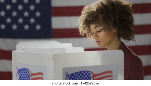 MCU Profile Young, Mixed Race Woman, Alone Casting Vote At Polling Station. She Looks Down At Ballot Paper, Out Of Frame. US Flag In Background.