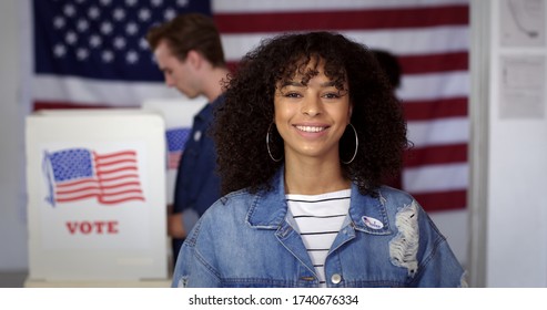 MCU Young Hispanic woman in denim jacket with a new "I Voted" sticker, smiling and standing in front of voters in polling booths with US flag in background