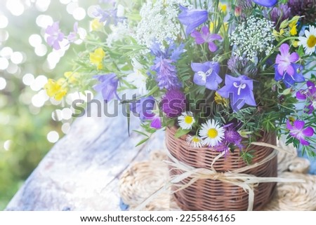 Meadow flowers bouquet in wicker mug on blue vintage wooden background outdoors in natural morning light, vivid wild flowers in background of sparkling water in summertime, close up view