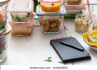 Meal prep scene with piles of glass jars full of homemade meal. A black notepad with a pen on it is in the front. - Shutterstock ID 1618634032