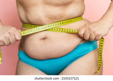 Measure female waist with tape closeup, folds on stomach, loose skin, fat and cellulite. Naked overweight plus size girl on pink background in blue underwear. Concept of dieting and body control.