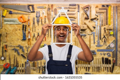 measurement, construction and building - happy smiling indian worker or builder in helmet with folding ruler in shape of home over working tools on wall at workshop background