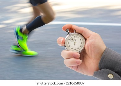Measuring The Running Speed Of An Athlete Using A Mechanical Stopwatch. Hand With A Stopwatch On The Background Of The Legs Of A Runner