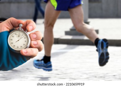 Measuring The Running Speed Of An Athlete Using A Mechanical Stopwatch. Hand With A Stopwatch On The Background Of The Legs Of A Runner