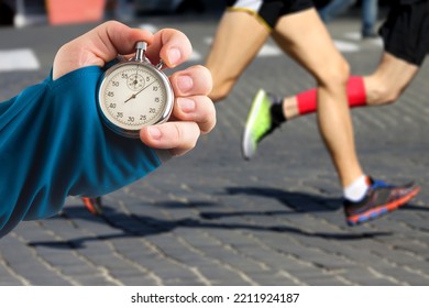 Measuring The Running Speed Of An Athlete Using A Mechanical Stopwatch. Hand With A Stopwatch On The Background Of The Legs Of A Runner.
