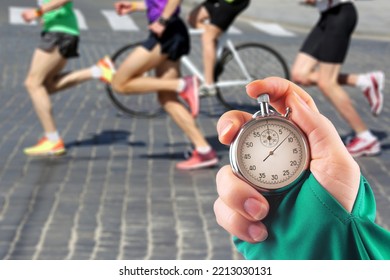 Measuring The Running Speed Of An Athlete Using A Mechanical Stopwatch. Hand With A Stopwatch On The Background Of The Legs Of A Runner.
