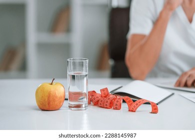 Measuring tape, an apple and glass of water are on the table. Young female doctor in white coat is indoors.