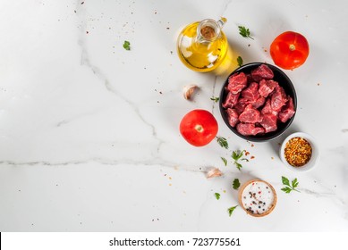 Meat, beef. Fresh raw chopped goulash, beef cubes in a bowl. Spices (salt, pepper), tomatoes, garlic, onions. On a white marble table, with a fork for meat and a knife. Top view copy space