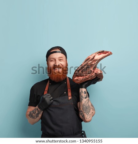 Meat consumption concept. Satisfied man meatpaking worker, smiles gladfully, carries raw pork on bone, works on slaughthouse, keeps hand on apron, wears protective gloves, isolated on blue wall