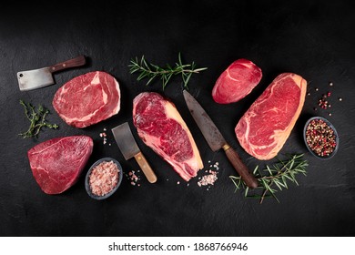 Meat flat lay. Various cuts of meat, overhead flat lay shot with knives, salt, pepper, and herbs, on a black slate background