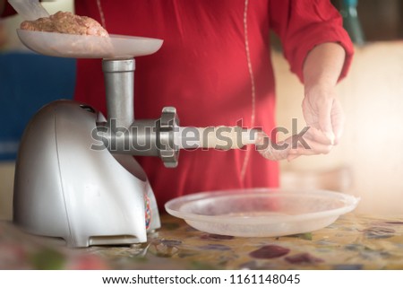 Meat grinder with fresh forcemeat and woman making sausages in kitchen