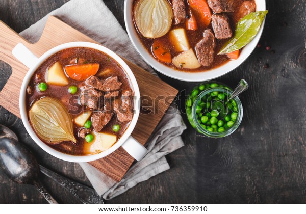 Meat stew with beef, potato,\
carrot, pepper, spices, green peas. Slow cooked meat stew, bowl,\
wooden background. Hot autumn/winter dish. Closeup. Top view.\
Comfort food. Homemade\
soup/ragout/casserole
