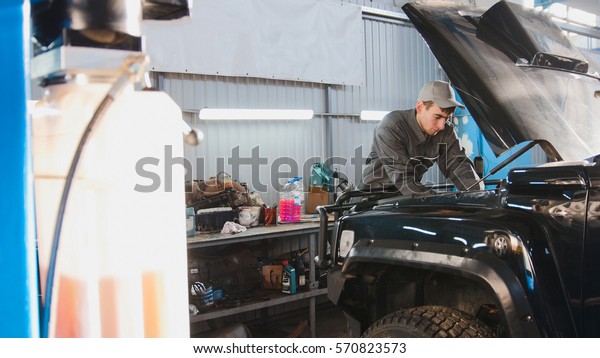 Mechanic in automobile garage checking hood of\
the car, wide angle,\
horizontal