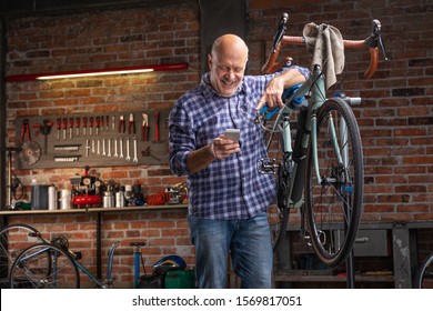 Mechanic In A Bicycle Repair Shop Using His Mobile Phone Reading A Text Message With A Smile As He Leans On The Bike He Is Working On